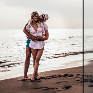 May include: A person with long blonde hair wearing a white button-down shirt over a pink bikini top and blue shorts, embraced by another person wearing a white t-shirt, blue shorts, and a white fedora hat while standing on a sandy beach.