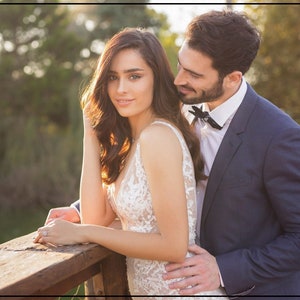 May include: A white sleeveless gown with intricate floral lace embroidery and a deep V-neckline, paired with a dark navy blue suit jacket, a crisp white collared shirt, and a small black bow tie, worn by two individuals standing together.