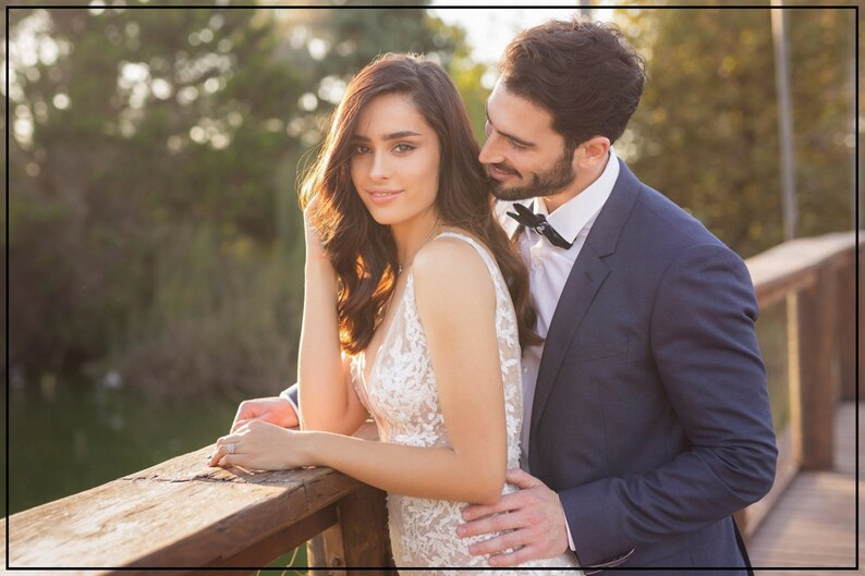 May include: A white sleeveless gown with intricate floral lace embroidery and a deep V-neckline, paired with a dark navy blue suit jacket, a crisp white collared shirt, and a small black bow tie, worn by two individuals standing together.
