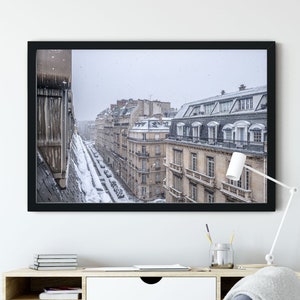 May include: A black framed photograph of a Parisian street scene with snow falling. The buildings are tall and narrow, with many windows. The street is lined with parked cars and a few people walking.