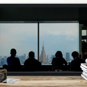May include: Silhouettes of four people looking out a large window at a city skyline. The city is in the distance and the people are in the foreground. The window is framed by a black frame.