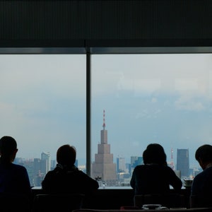 May include: Silhouettes of four people sitting in front of a large window with a city skyline view. The window is divided into four panes. The people are facing the window and appear to be looking out at the city.