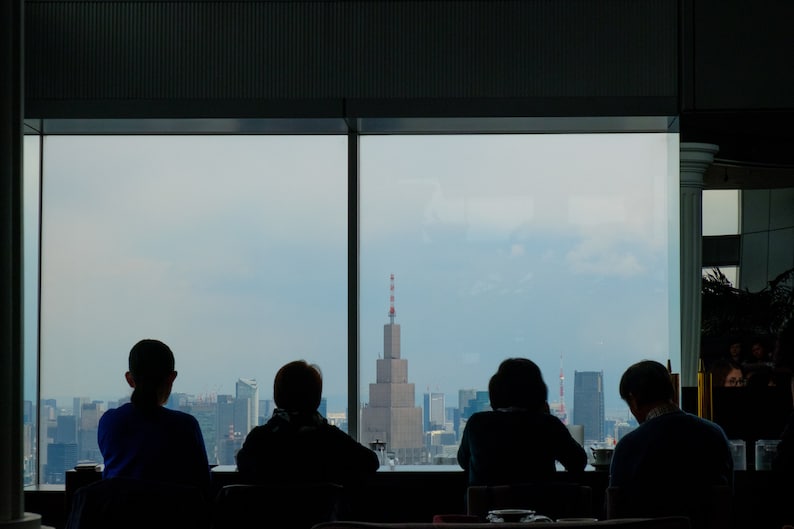 May include: Silhouettes of four people sitting in front of a large window with a city skyline view. The window is divided into four panes. The people are facing the window and appear to be looking out at the city.