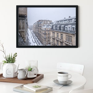 May include: A black framed photograph of a Parisian street scene with snow falling. The buildings are tall and narrow, with many windows. The street is lined with parked cars and a few pedestrians.