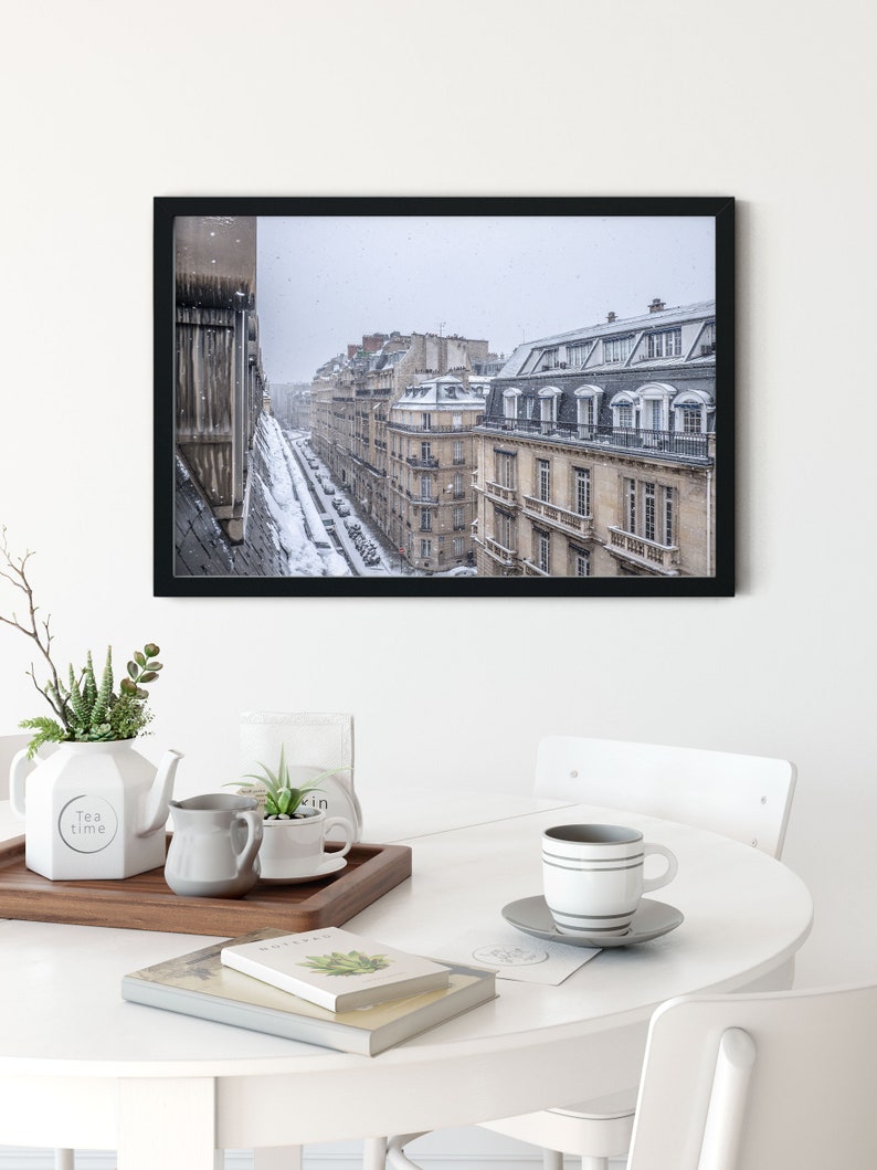 May include: A black framed photograph of a Parisian street scene with snow falling. The buildings are tall and narrow, with many windows. The street is lined with parked cars and a few pedestrians.