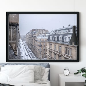 May include: A framed photograph of a snowy Parisian street scene. The photo shows a narrow street lined with buildings, with snow falling gently. The buildings are all in a similar style, with tall windows and ornate details. The street is empty, except for a few parked cars.
