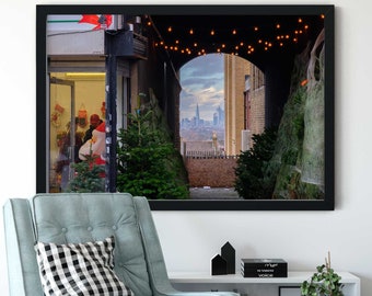 May include: A framed photograph of a narrow alleyway decorated with Christmas lights, leading to a view of a city skyline. The alleyway is lined with potted Christmas trees and a shop window is visible on the left.