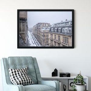 May include: A black framed photograph of a snowy Parisian street scene. The buildings are tall and narrow, with many windows. The street is lined with cars and trees. The snow is falling softly, creating a peaceful atmosphere.