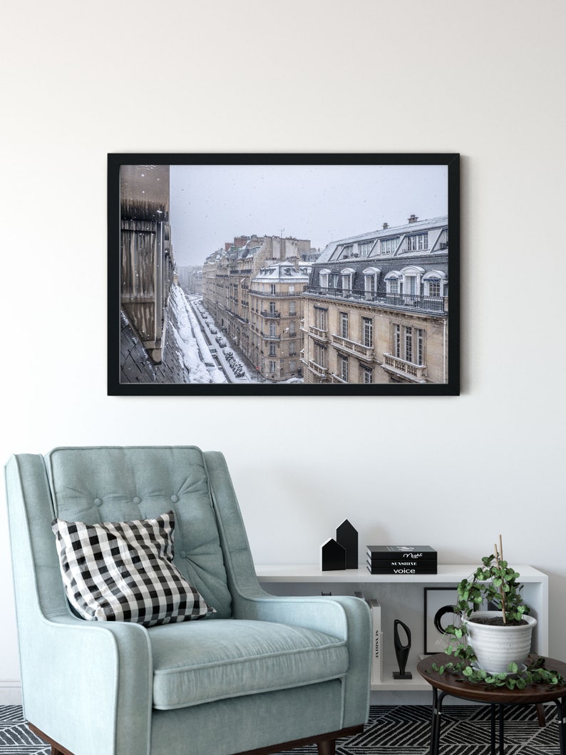 May include: A black framed photograph of a snowy Parisian street scene. The buildings are tall and narrow, with many windows. The street is lined with cars and trees. The snow is falling softly, creating a peaceful atmosphere.