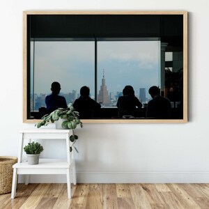 May include: A framed photograph of four people sitting in front of a large window with a view of a city skyline. The people are silhouetted against the window, and the city skyline is visible in the background.