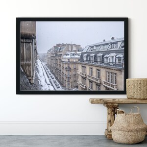May include: A framed photograph of a Parisian street scene with snow falling. The buildings are tall and narrow, with many windows. The street is lined with parked cars. The photo is in a black frame.