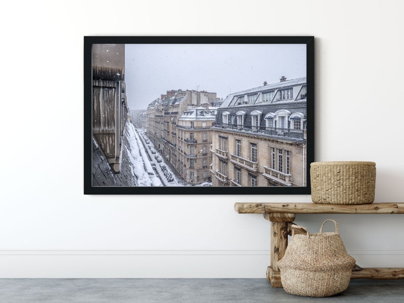 May include: A framed photograph of a Parisian street scene with snow falling. The buildings are tall and narrow, with many windows. The street is lined with parked cars. The photo is in a black frame.