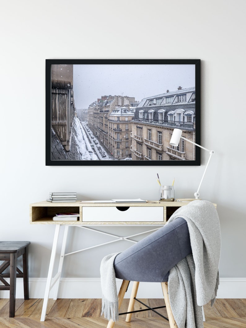 May include: A black framed photograph of a Parisian street scene with snow falling. The buildings are tall and narrow, with many windows. The street is lined with parked cars and a few people walking.