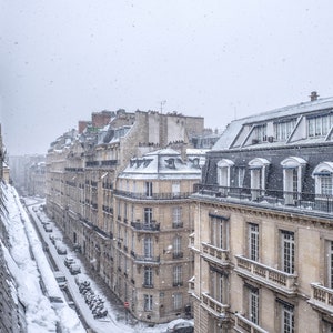 May include: A snowy Parisian street scene with a view of multiple buildings. The buildings are all made of stone and have a traditional French architecture. The street is covered in snow and there are cars parked on the side of the street.