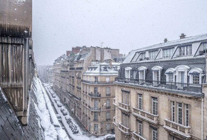 May include: A snowy Parisian street scene with a view of multiple buildings. The buildings are all made of stone and have a traditional French architecture. The street is covered in snow and there are cars parked on the side of the street.