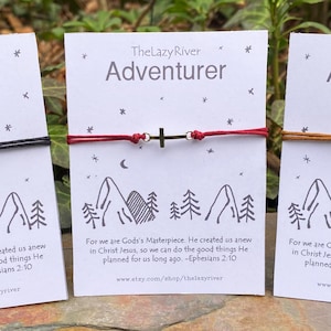 May include: Three bracelets with a cross charm on a white card with the text "The Lazy River Adventurer" and a mountain scene. The bracelets are black, red, and brown.