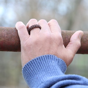 May include: A close-up of a hand wearing a brown braided ring. The hand is resting on a rusty metal bar.