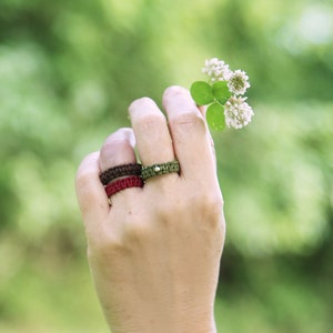 May include: A hand wearing three rings, one brown, one red, and one green, with a small white stone on the green ring. The hand is holding a small white flower with green leaves.