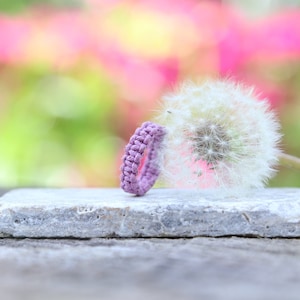 May include: A purple braided ring sits on a gray stone next to a white dandelion.