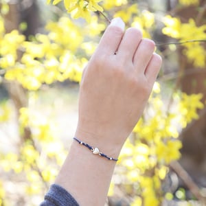 Puede incluir: Un brazalete de cuerda azul marino con un dije de corazón plateado. El brazalete se lleva en la muñeca de una persona sobre un fondo de flores amarillas.