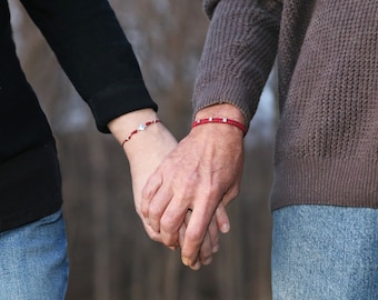 Puede incluir: Una pareja se toma de la mano, cada uno lleva una pulsera de cuerda roja con abalorios plateados.