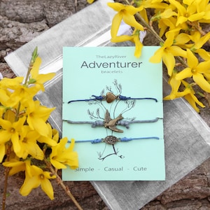 May include: Three blue and gray friendship bracelets with charms, including a heart, a bird, and a circle. The bracelets are packaged in a clear bag with a green card that says "The Lazy River Adventurer bracelets" and "Simple - Casual - Cute".