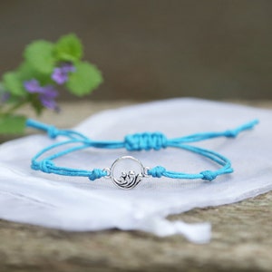 Puede incluir: Un brazalete de amistad azul turquesa con un dije de plata con diseño de ola. El brazalete está atado con un nudo y está sobre una superficie de tela blanca.