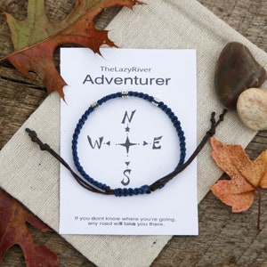May include: A navy blue braided bracelet with silver beads and a compass design. The bracelet is on a white card with the text "TheLazyRiver Adventurer" and "If you don't know where you're going, any road will take you there."