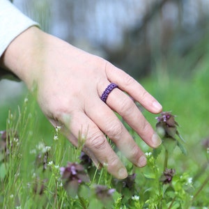 May include: A hand wearing a purple braided ring reaches out to touch a patch of green grass with small white flowers.