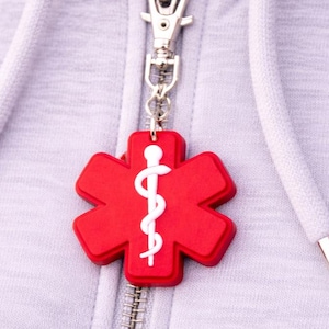Op de afbeelding: Een rode medische symboolbedel met een witte staf van Asclepius hangt aan een zilveren rits. De bedel is bevestigd aan een lichtgrijze hoodie met witte trekkoorden en zilveren uiteinden.