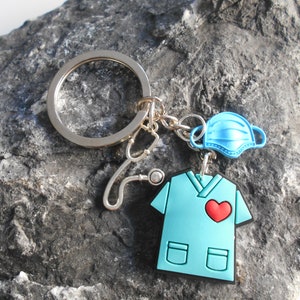 May include: A blue scrubs keychain with a red heart, a stethoscope, and a blue face mask. The keychain is attached to a silver key ring.