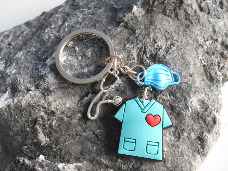 May include: A blue scrubs keychain with a red heart, a stethoscope, and a blue face mask. The keychain is attached to a silver key ring.