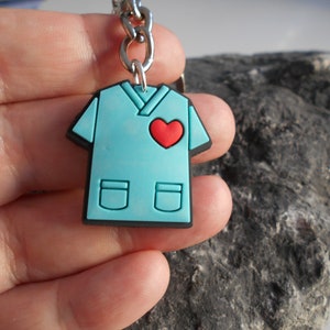 May include: A blue scrub top keychain with a red heart on the front. The keychain is made of rubber and has a silver metal chain.