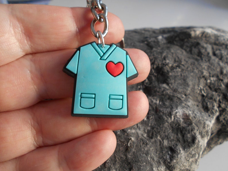May include: A blue scrub top keychain with a red heart on the front. The keychain is made of rubber and has a silver metal chain.