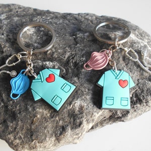 May include: Two keychains with a blue and pink medical mask, a stethoscope, and a blue scrub top with a red heart on the front.