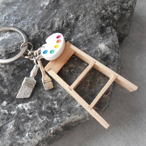 Op de afbeelding: Een zilveren sleutelhanger met miniatuur bedels met schilderthema. Het bevat een wit verfpalet met gekleurde verfvlekken, twee penselen en een kleine houten ladder. De sleutelhanger is geplaatst tegen een grijze stenen achtergrond.