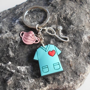 May include: A blue scrubs keychain with a red heart, a pink face mask, and a silver stethoscope charm.