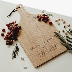 Könnte beinhalten: Ein personalisiertes Holzbrett zum Schneiden mit den Namen "Verena" und "Simon", die zusammen mit einem Herz und einem floralen Design eingraviert sind. Das Brett ist von Trauben, Pistazien und grünen Zweigen umgeben.