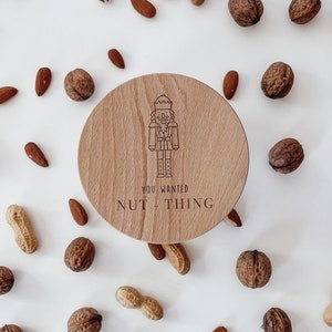 Op de afbeelding: Een ronde houten doos met een notenkrakerontwerp en de tekst "You wanted Nut-thing". De doos is omringd door noten en pinda's op een witte achtergrond.
