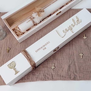 Könnte beinhalten: Eine weiße Holzkiste mit einer langen weißen Kerze, verziert mit Trockenblumen und Bindfaden. Die Kiste ist mit dem Namen "Leopold" und den Worten "Kommunionskerze von" beschriftet. Kleine goldene Kreuze sind verstreut.