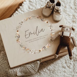Pode incluir: Uma caixa de memória de madeira clara com o nome "Emilia" e um design de coroa floral. A caixa é acompanhada por um par de sapatos de bebé bege e um animal de pelúcia castanho. A caixa tem o texto "06.03.2026 | 22:04 Uhr | 3200 g | 42 cm" e "Ein Stück Kindheit für immer".
