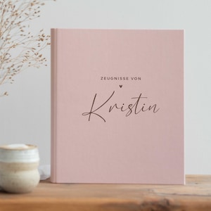 Puede incluir: Un libro de tapa dura de color rosa claro con el texto "ZEUGNISSE VON" sobre un pequeño icono de corazón y el nombre "Kristin" en una fuente cursiva de color marrón oscuro. El libro está colocado en posición vertical sobre una superficie de madera.