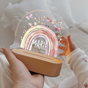 Può includere: Una targa semicircolare in acrilico trasparente inserita in una base di legno chiaro, con un arcobaleno ad acquerello in tenui tonalità pastello. Il nome MARIE e la data 18 august 2026 sono stampati in bianco, circondati da farfalle colorate.
