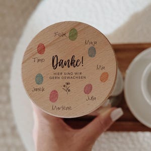 Puede incluir: Una tapa de madera con la palabra alemana "Danke!" (Gracias) y la frase "Hier sind wir gern gewachsen" (Aquí hemos crecido) se sostiene sobre una taza y un platillo blancos. La tapa tiene puntos de colores y nombres. Se ve una muñeca con pulseras doradas y un tatuaje.