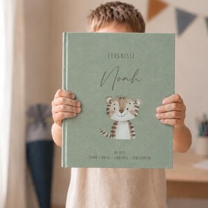 Puede incluir: Un libro de tapa dura color verde salvia con textura de tela que presenta el texto ZEUGNISSE, el nombre Noah y una ilustración de un cachorro de tigre. Debajo del tigre, el texto dice DU BIST STARK MUTIG LIEBEVOLL EINZIGARTIG en gris oscuro.