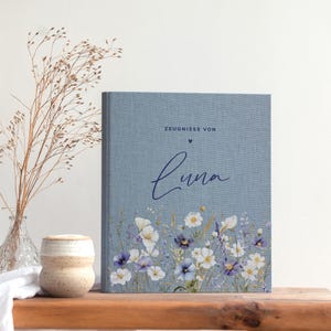 Könnte beinhalten: Ein blaues, stoffgebundenes Buch mit strukturierter Oberfläche und einem Blumendruck aus weißen und violetten Wildblumen am unteren Rand. Der Text lautet "ZEUGNISSE VON" über einem kleinen Herz und dem Namen "Luna" in dunkelblauer Schreibschrift.