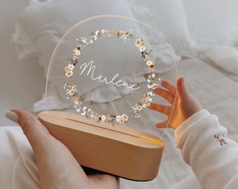 Puede incluir: Una luz de noche personalizada con una base de madera y una parte superior de acrílico transparente y circular. El acrílico presenta un diseño de corona floral y el nombre "Marlene" en escritura cursiva. La luz es sostenida por manos.