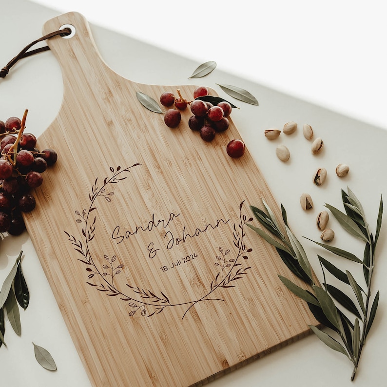Puede incluir: Una tabla de cortar de bamb&uacute; personalizada con un dise&ntilde;o de corona floral y los nombres "Sandra & Johann" y la fecha "18. Juli 2024" grabados en color burdeos.