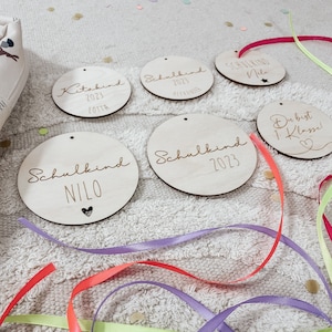 Op de afbeelding: Vijf ronde houten ornamenten met de tekst "Schulkind" (Duits voor "schoolkind") en het jaar "2023" erop gegraveerd. Eén ornament heeft de tekst "Kitakind" (Duits voor "kleuterkind") en het jaar "2023". Eén ornament heeft de tekst "Du bist 1. Klasse!" (Duits voor "Je bent in de 1e klas!").