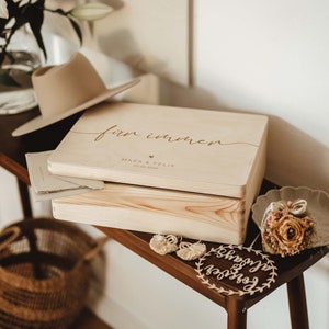 Op de afbeelding: Een houten doos met de inscriptie "für immer" (Duits voor "voor altijd") en de namen "Mara & Felix" en de datum "02.08.2023". De doos staat op een houten tafel met een klein houten bord waarop "happily ever after" staat.